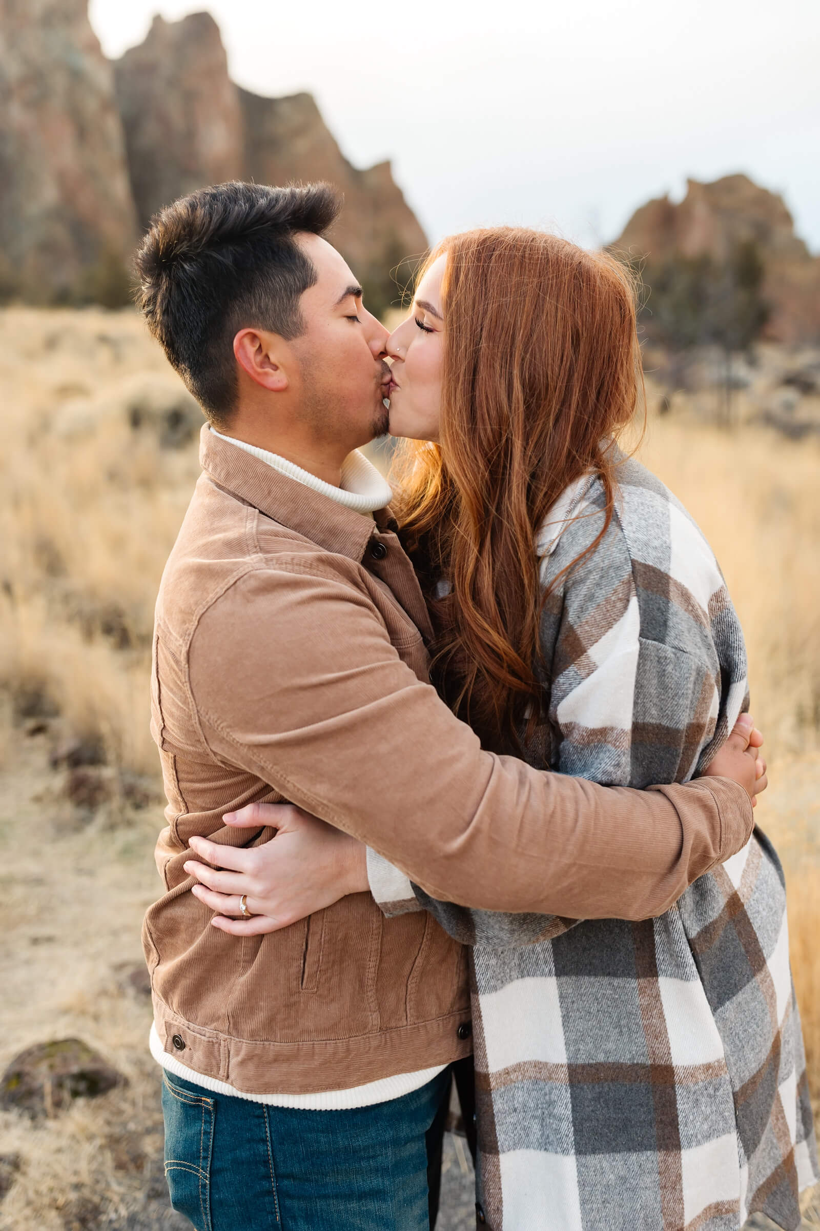 dark haired male in leather jacket and woman with red hair and gray plaid shacket hugging and kissing during smith rock family photoshoot