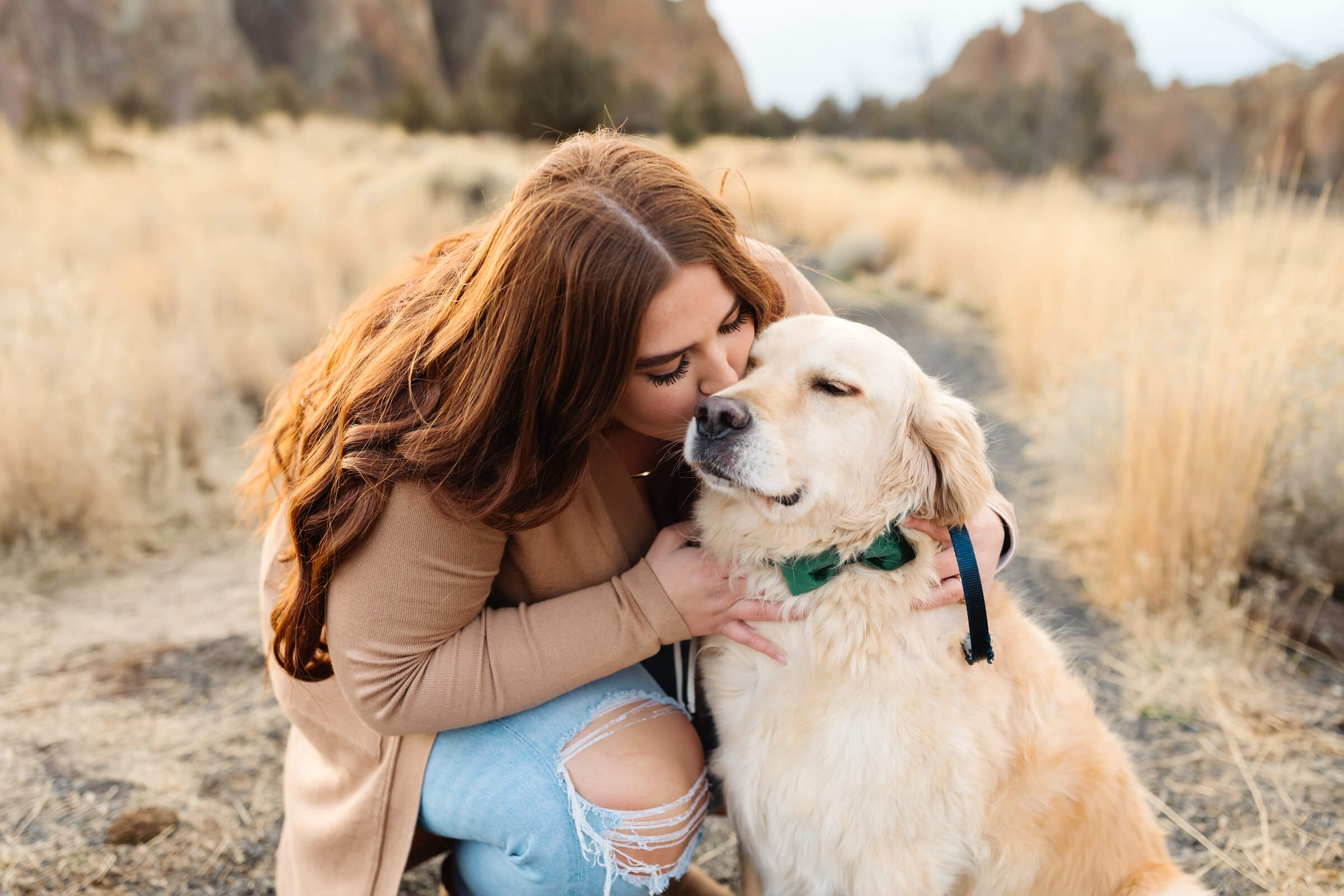 woman with red hair and tan long sweater kneeling and kissing golden retriever with dark green bow tie during smith rock family photoshoot