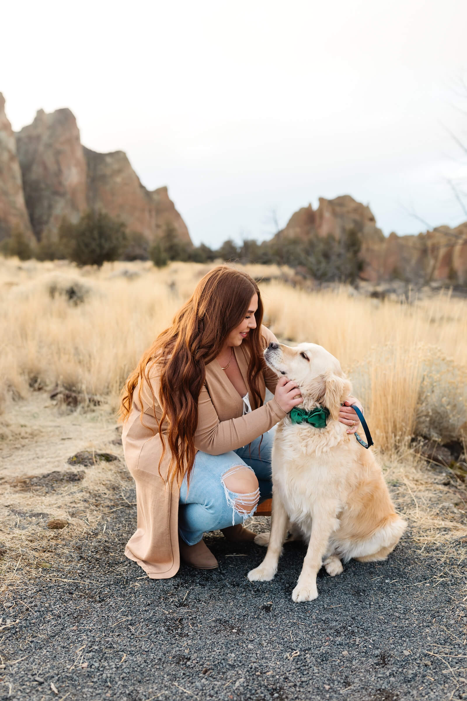 woman with red hair and tan long sweater kneeling and looking at golden retriever with dark green bow tie during smith rock family photoshoot