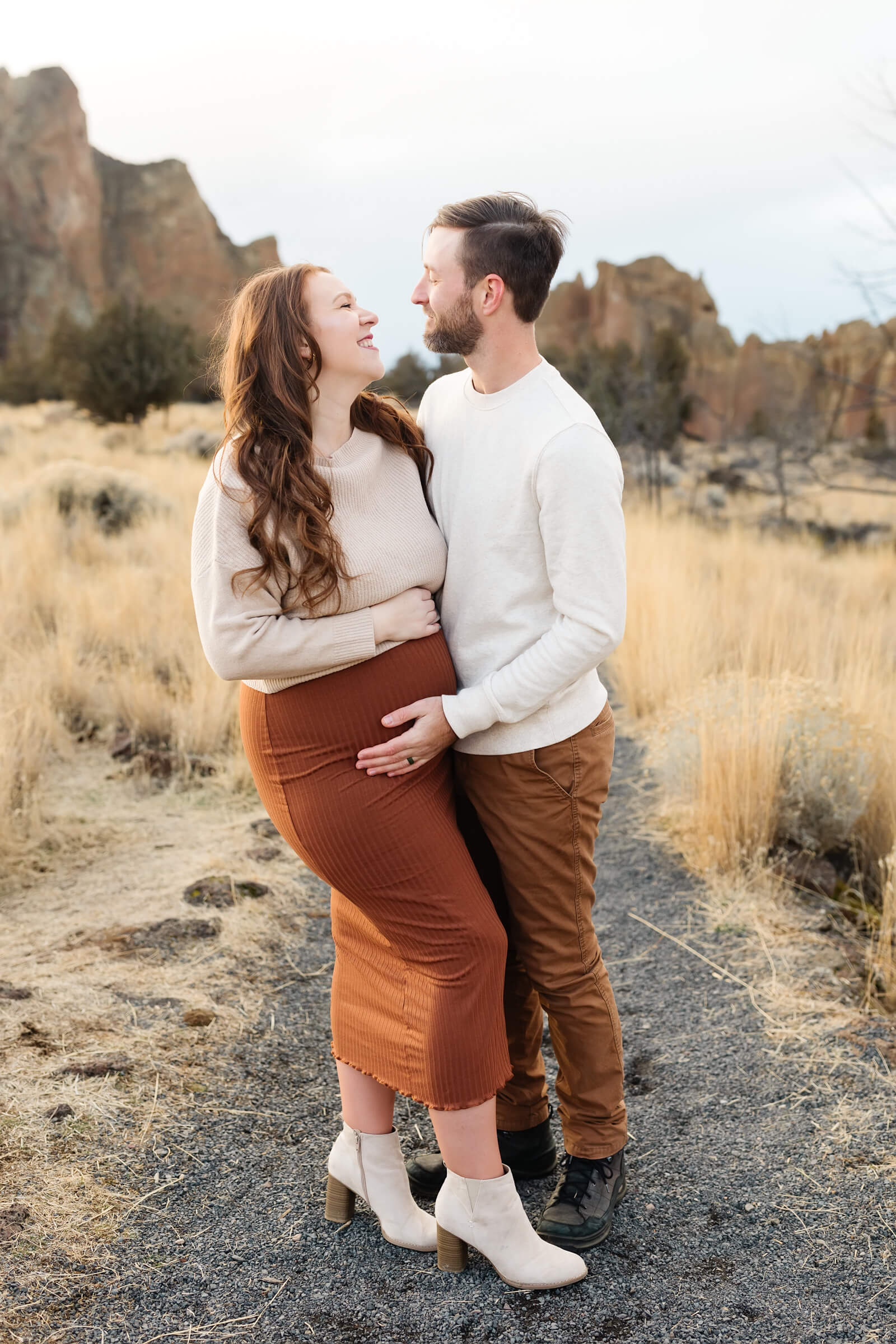 expecting mother with red hair and wearing terracotta skirt smiling at husband touching her pregnant belly at smith rock