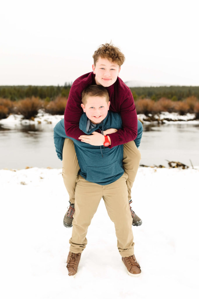 older brother in maroon sweater piggy backing younger brother in blue sweater next to Deschutes River in Sunriver Oregon during family photoshoot