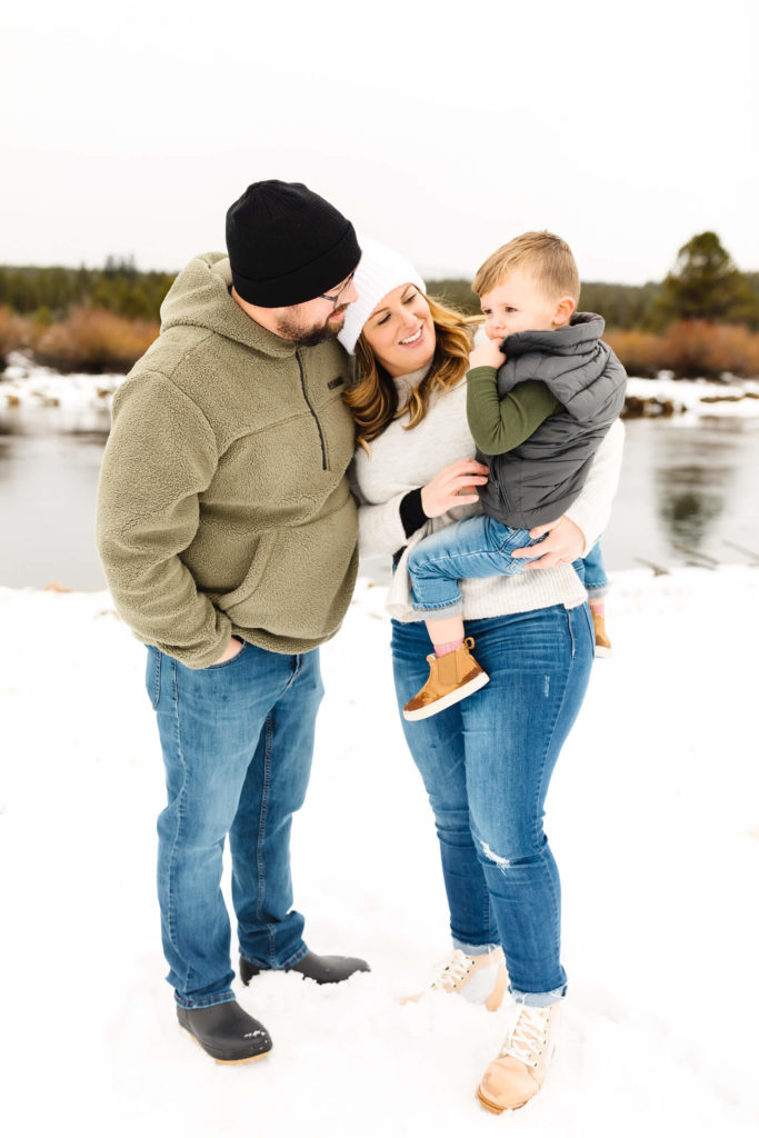 mother in white sweater and white pom pom beanie holding little boy while father smiles at them in Sunriver snow family photoshoot 