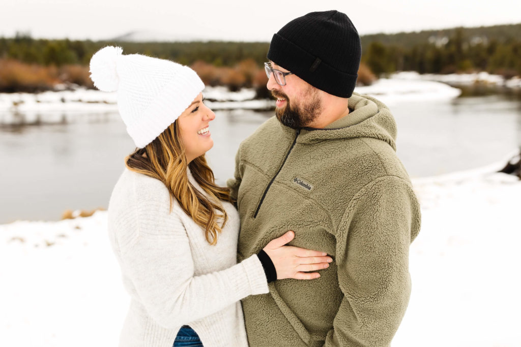 woman in white sweater and pom pom beanie and male in green fleece jacket standing in snow next to Deschutes river in Sunriver during family photoshoot