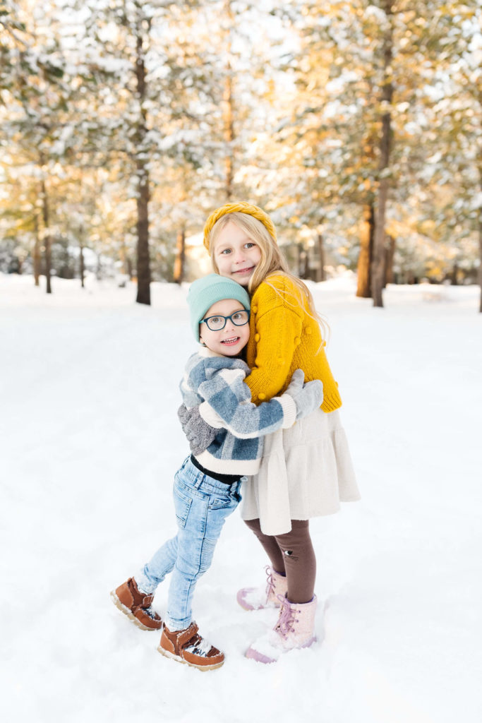 blonde brother and sister in yellow sweater hugging in the snow during winter family photoshoot