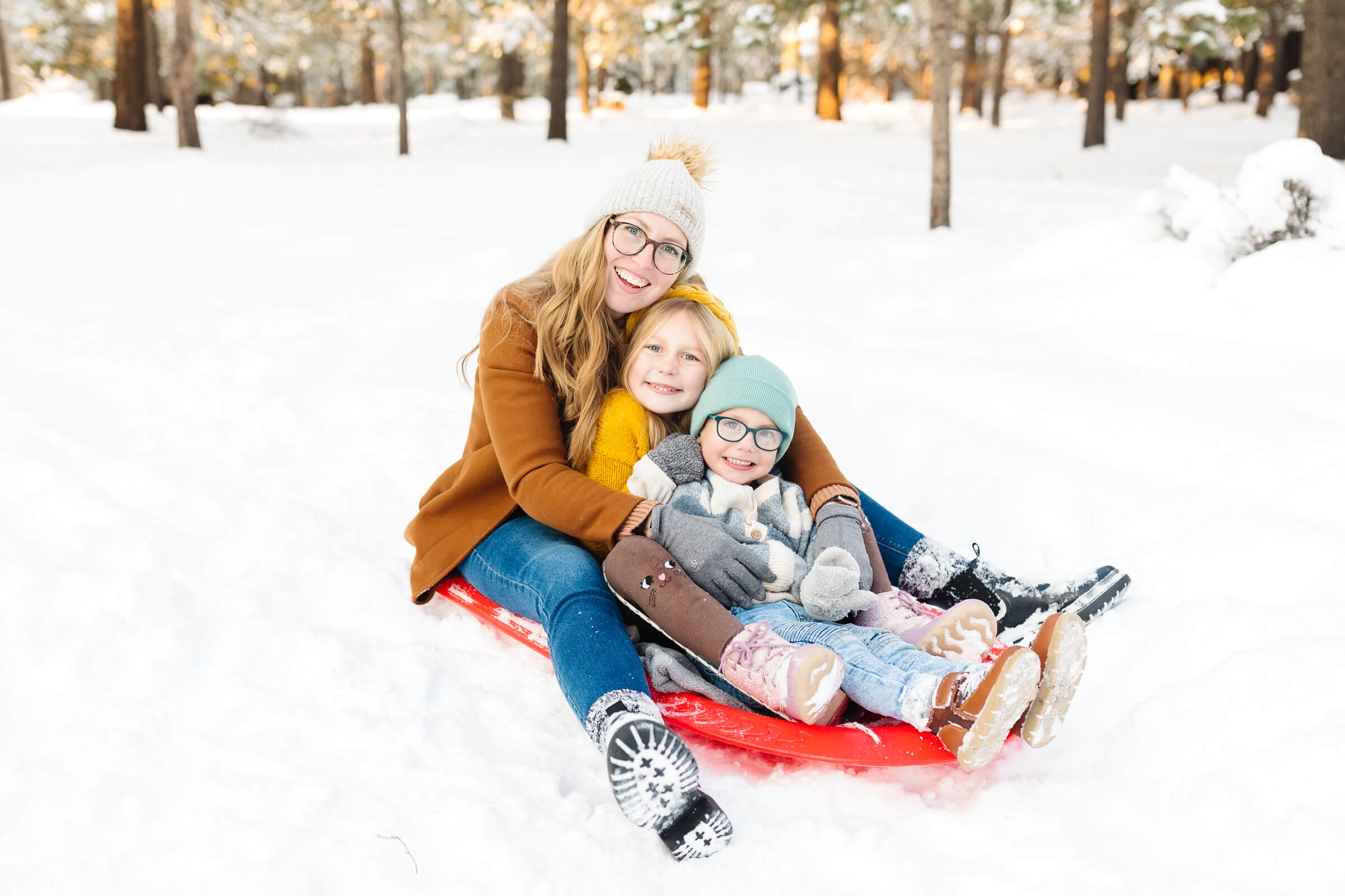blonde mother and two children sitting in a red sled in the snow during winter family photoshoot