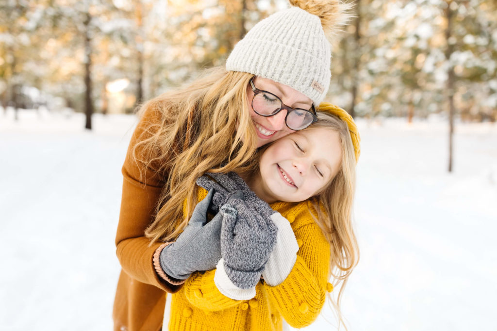 blonde mother in brown jacket hugging young daughter in yellow sweater during winter family photoshoot