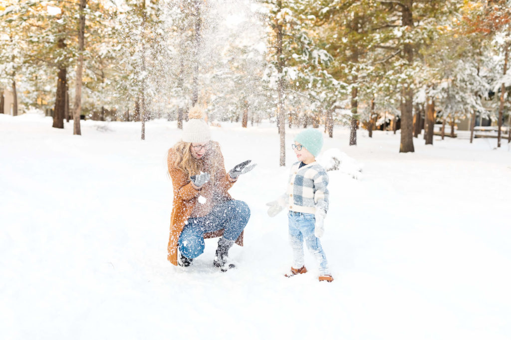 mother in brown wool jacket throwing snow with her son during winter family photoshoot