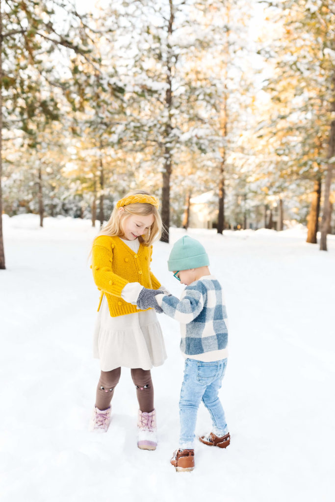 blonde little girl in yellow sweater and blonde little boy in blue plaid sweater playing ring around the rosie in the snow during winter family photoshoot