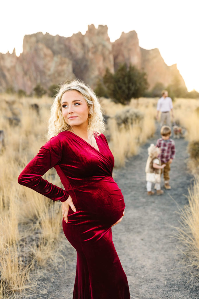 blonde woman in red velvet dress holding pregnant belly at maternity family photoshoot while family stands in background