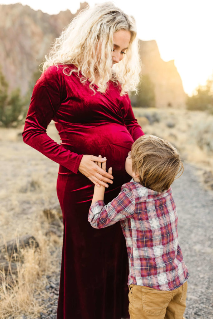 pregnant blonde woman in red velvet slit dress holding belly while little boy kisses her belly at smith rock