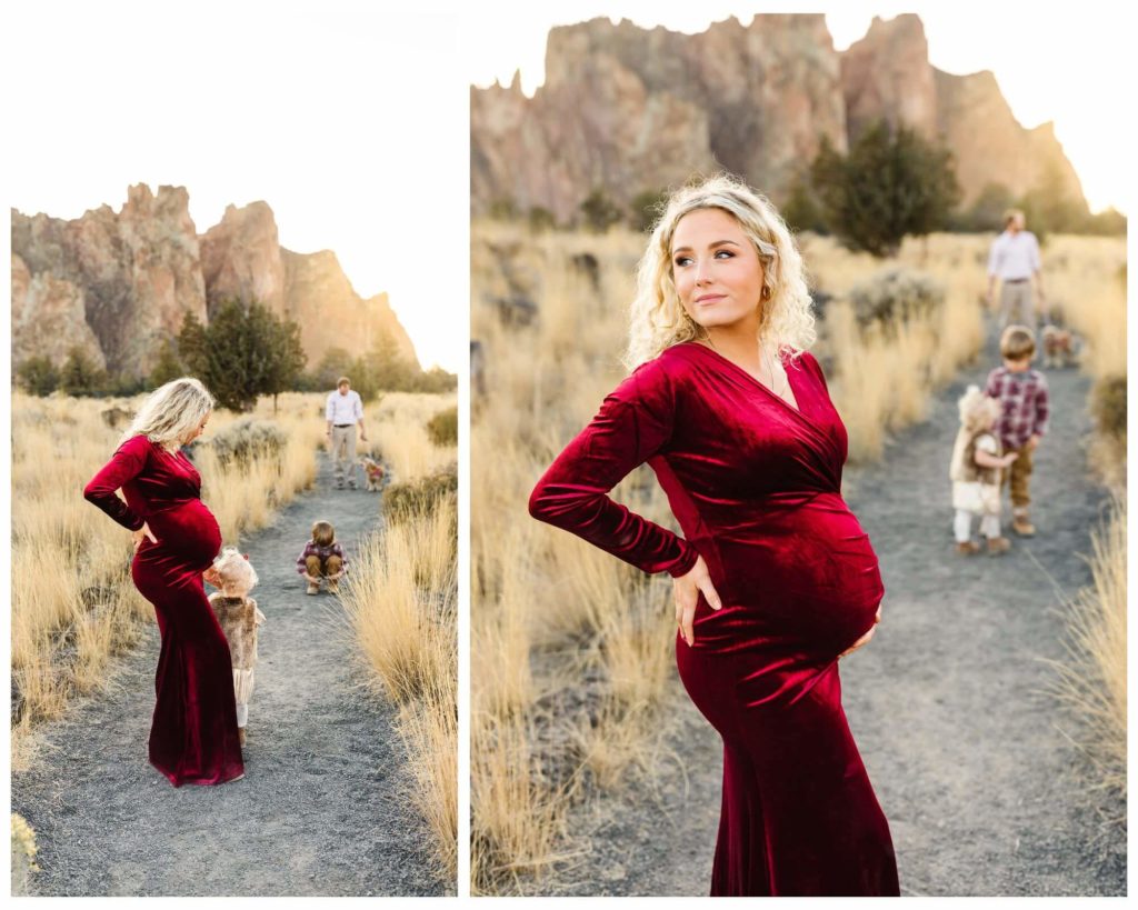 pregnant blonde woman in red velvet slit dress holding belly with smith rock and family in the background 