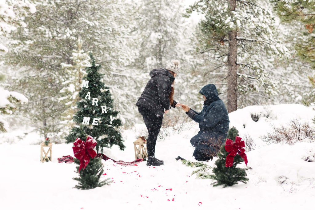 man in blue puffer coat kneeling on one knee and putting ring on womans finger during surprise Christmas proposal