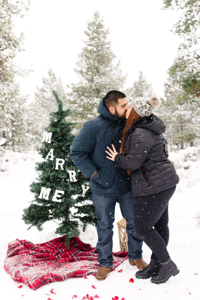 woman kissing man in front of christmas tree that says "Marry me"