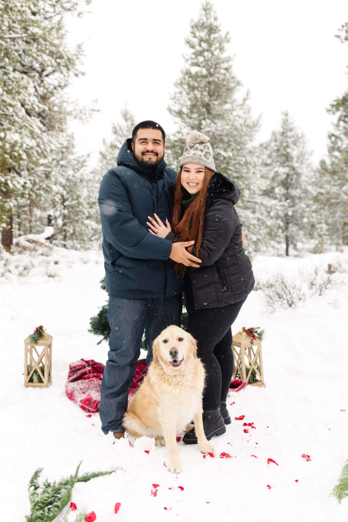 man in puffy blue jacket and woman with red hair with golden retriever in snow during christmas proposal