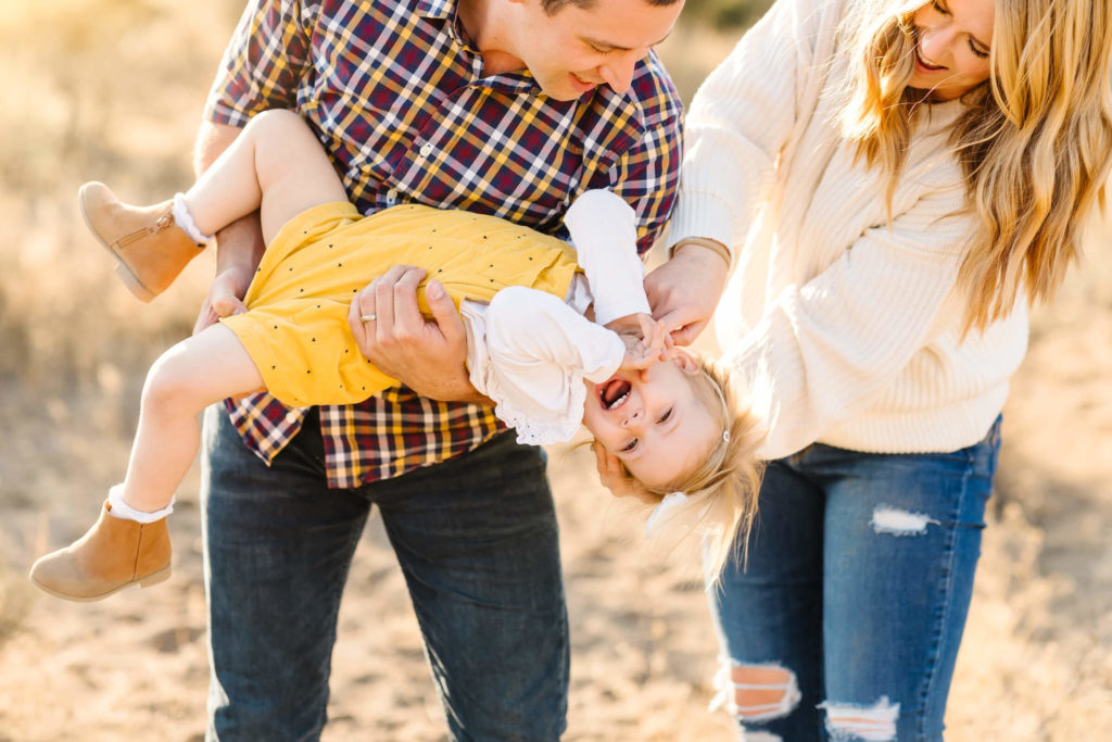 little blonde girl in yellow jumper being tickled by parents during family photoshoot at golden hour