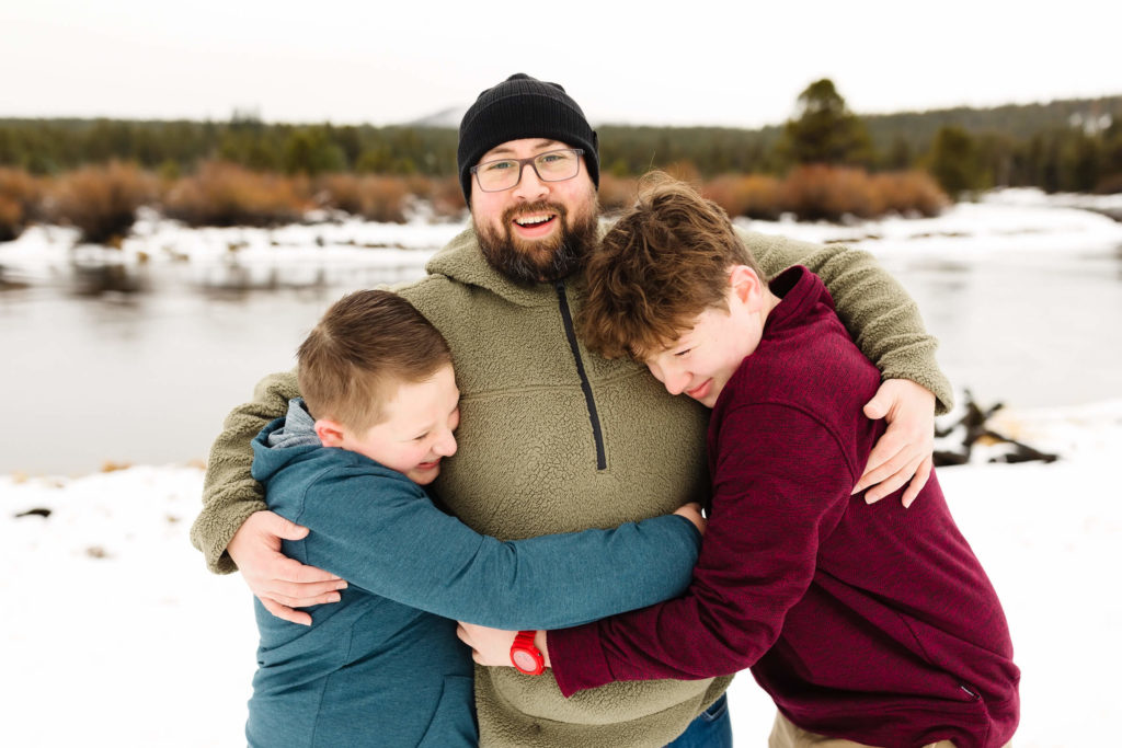 two young men hug their father near deschutes river during family photo session in snow