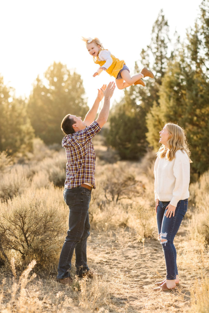 little blonde girl in yellow jumper being playfully thrown in air by father during golden hour cost of family photography session