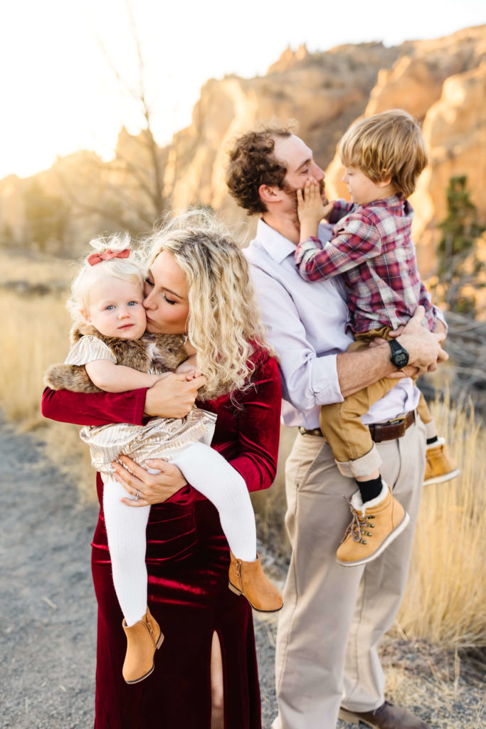 blonde mother in red velvet dress kissing young daughters cheek during family photoshoot