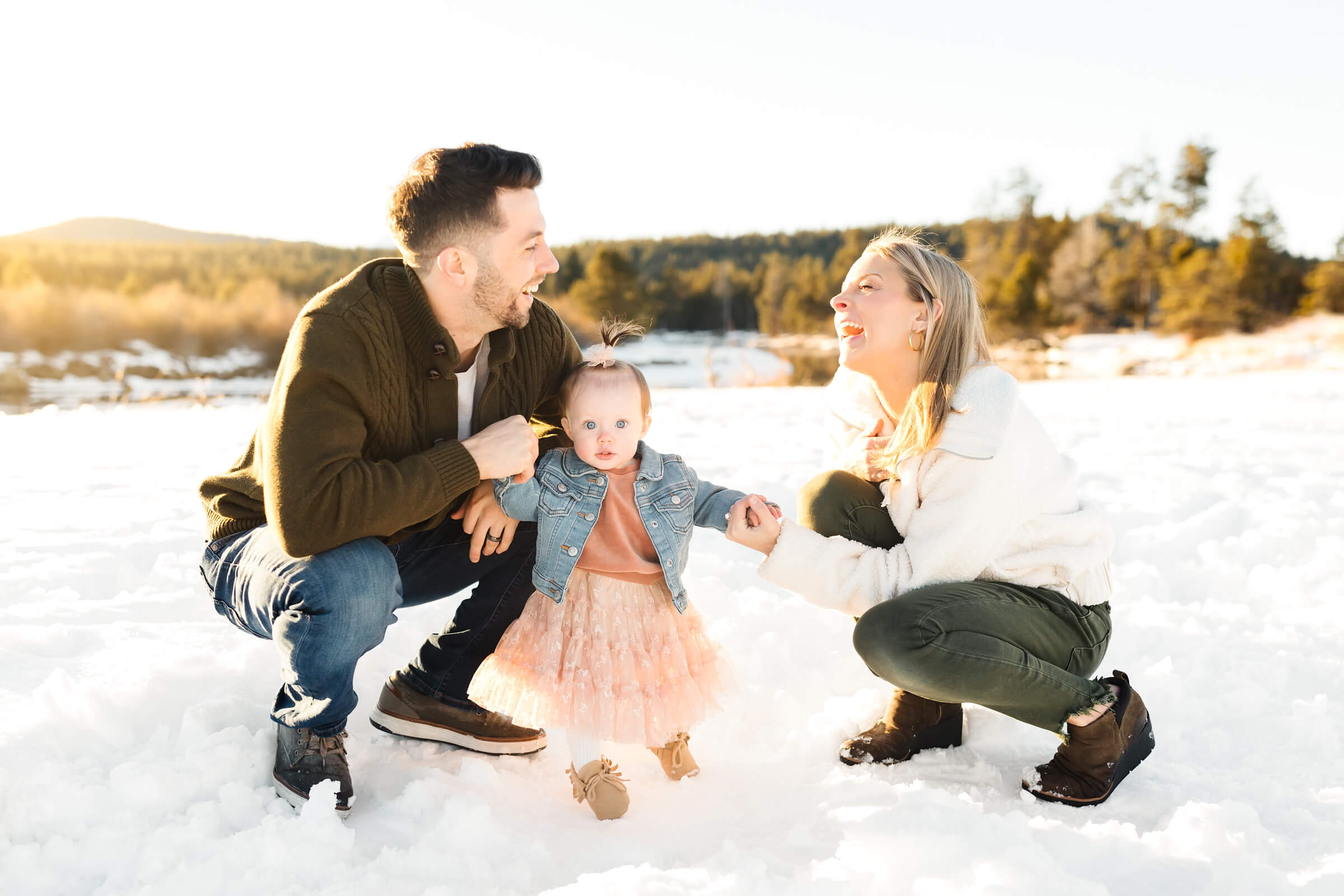 blonde mother and father kneeling in snow holding hands of 1 year old little girl in pink dress during joyful family photoshoot in bend or
