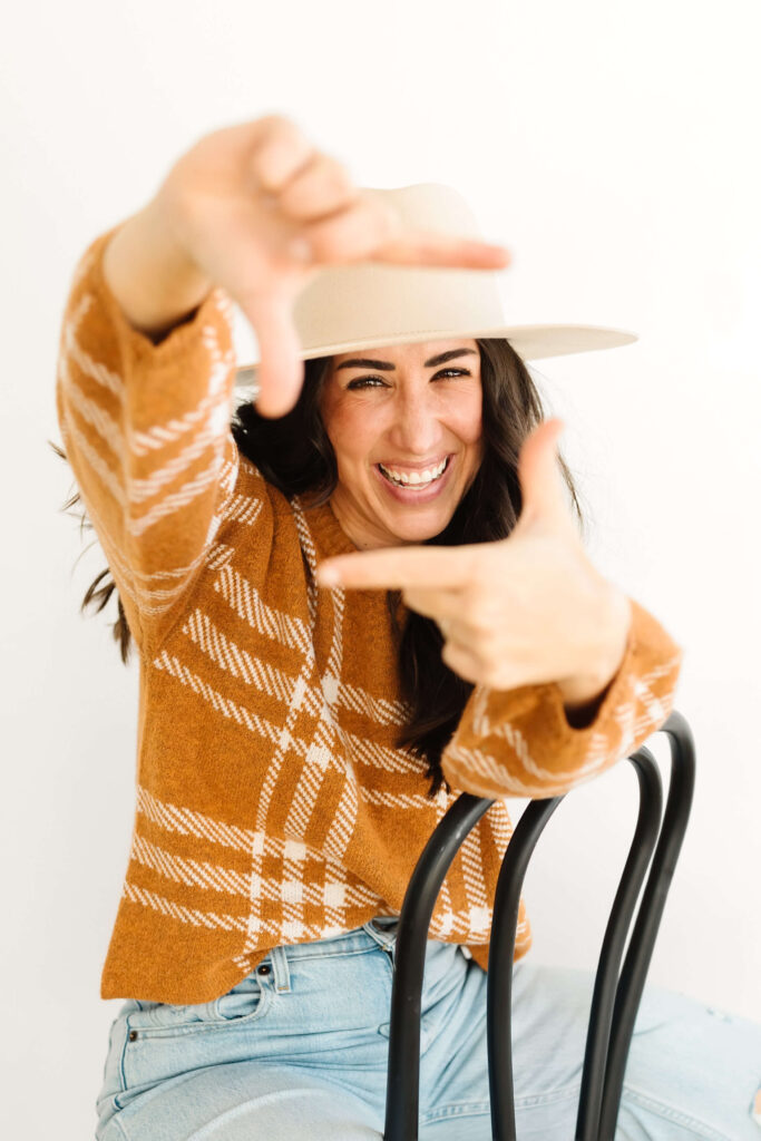 woman in orange sweater sitting backwards in black chair smiling through view finder fingers during creative poses for photographers headshots studio session