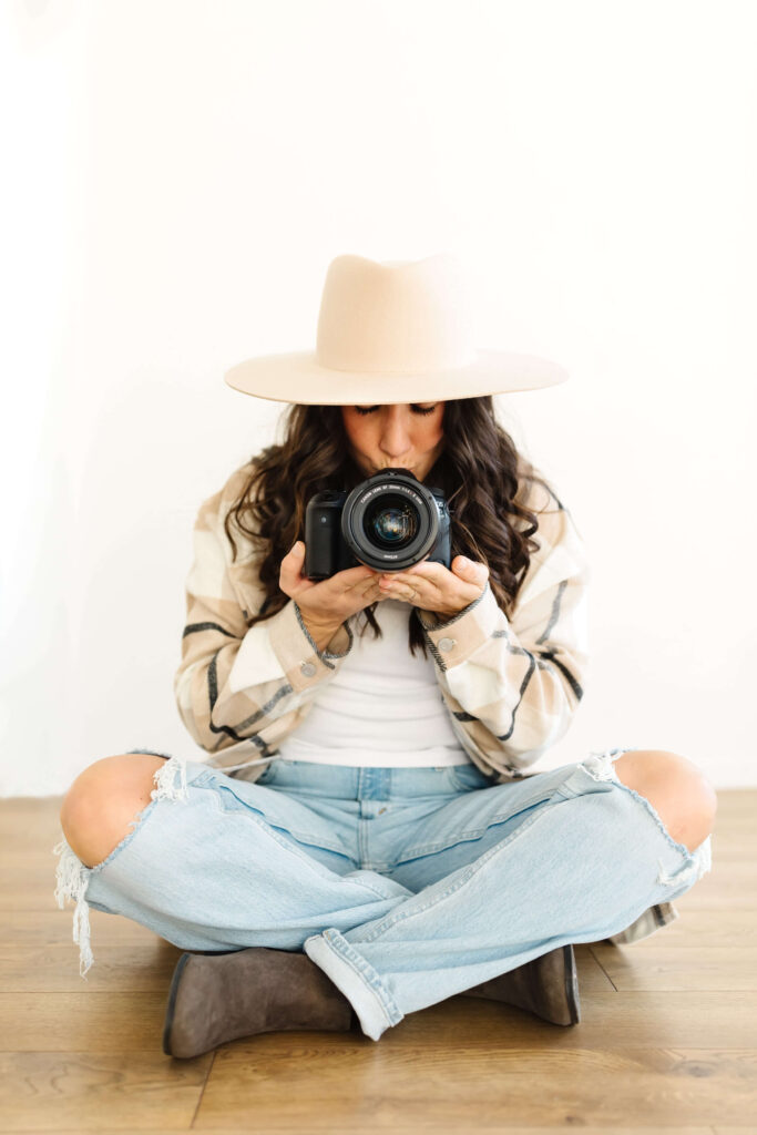 woman in wide brimmed hat sitting cross legged on floor kissing canon camera during creative poses photographers headshots session