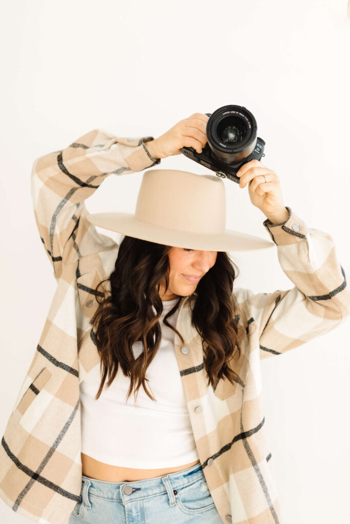 brunette in wide brimmed felt hat holding camera above her head during poses for creative photographers headshots session