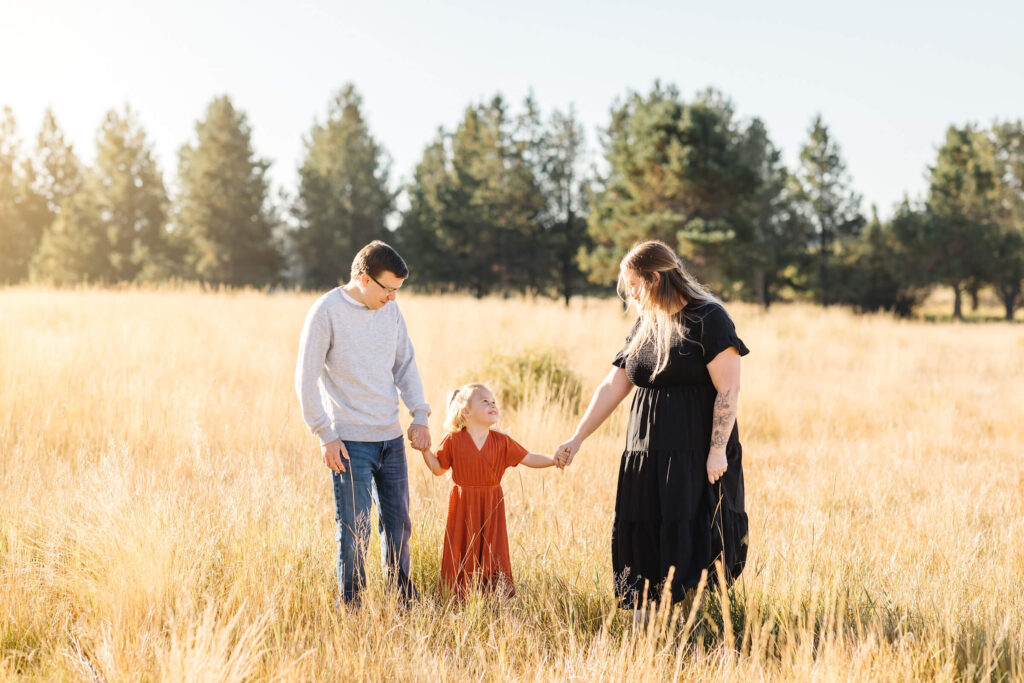 little blonde girl in orange dress standing between mother and father and holding their hands and looking up at her mother while working with Sunriver family photographers