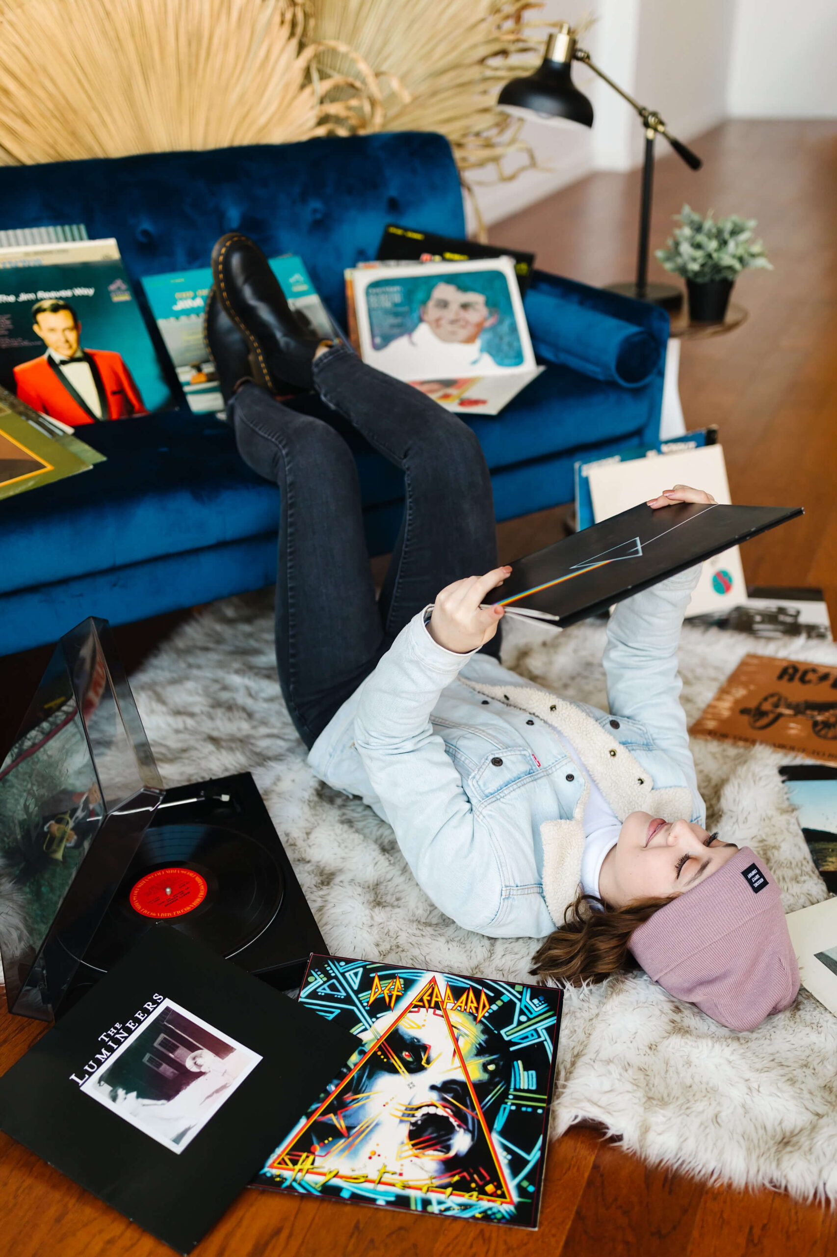 girl wearing pink beanie and blue jean jacket, laying on her back with feet up on blue velvet couch next to record player and vinyl record albums scattered around, during senior studio photoshoot