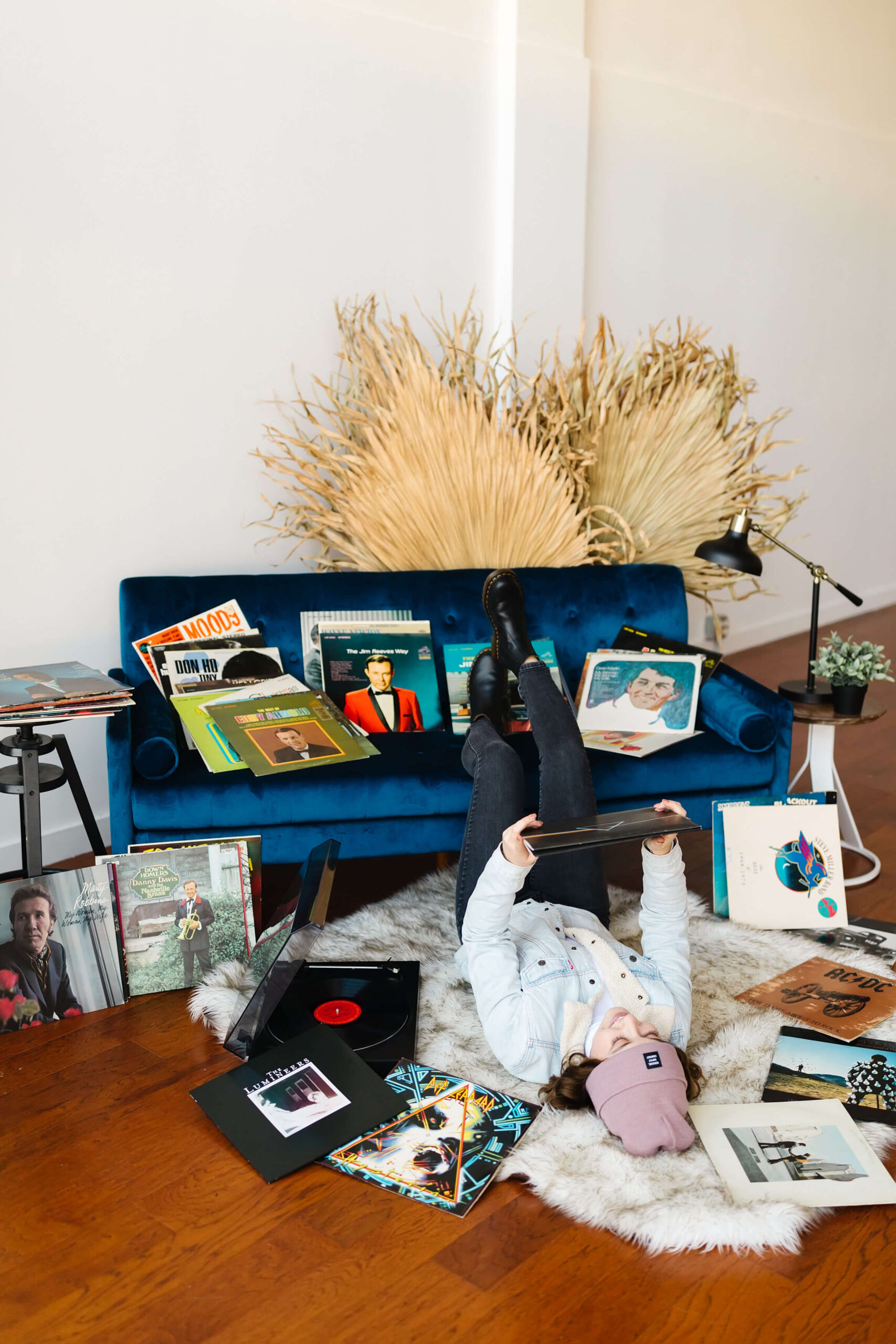 girl wearing pink beanie and blue jean jacket, laying on her back with feet up on blue velvet couch next to record player and vinyl record albums scattered around, during senior studio photoshoot