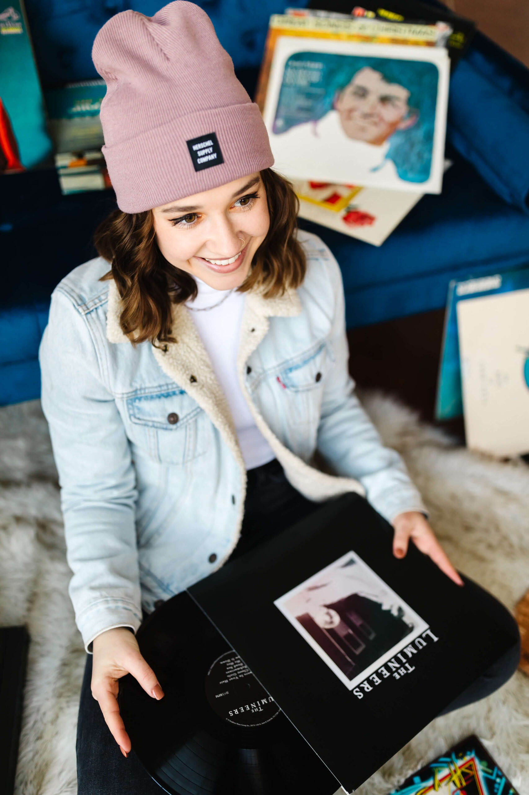 girl wearing pink beanie, blue jean jacket sitting in front of blue velvet couch taking a vinyl record album out of its cover during senior studio photoshoot