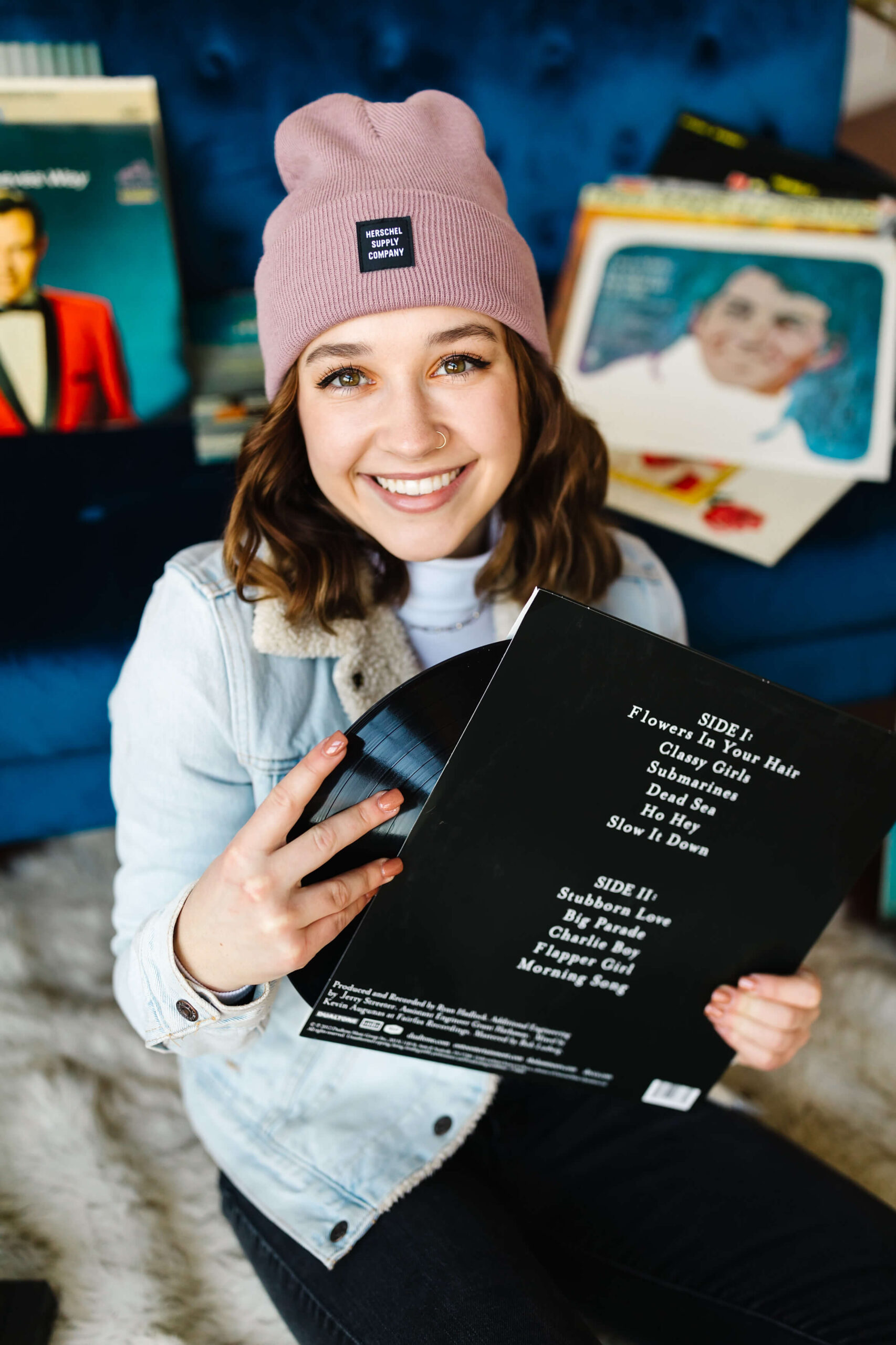 girl wearing pink beanie, blue jean jacket sitting in front of blue velvet couch taking a vinyl record album out of its cover during senior studio photoshoot