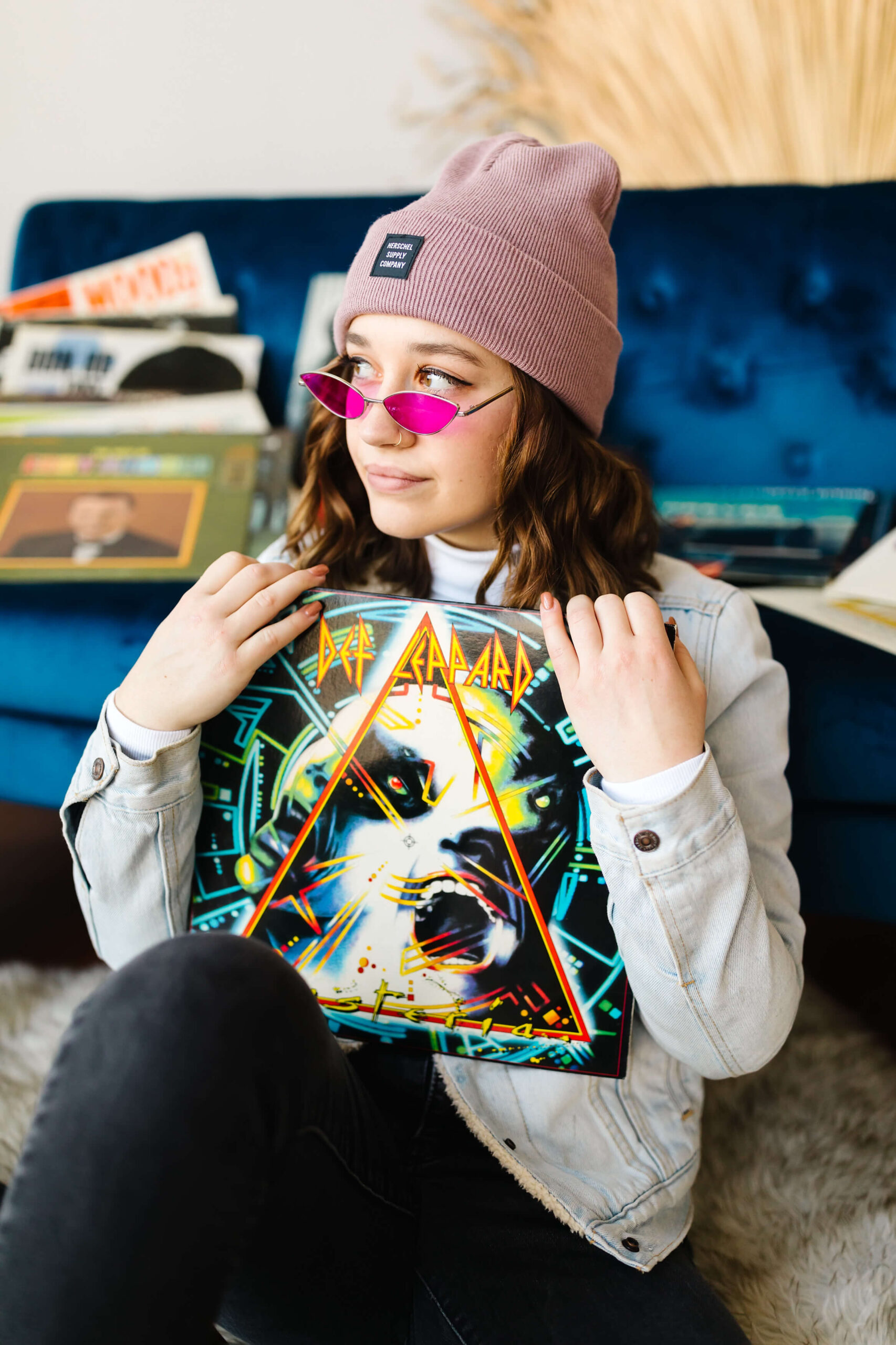 girl wearing pink beanie and blue jean jacket, leaning back on blue velvet couch next to record player and holding Def Leopard vinyl record with albums scattered around, during senior studio photoshoot