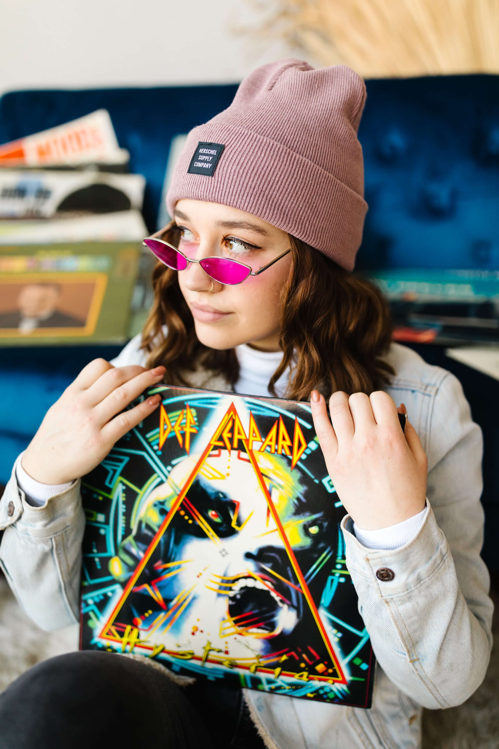 girl wearing pink beanie and blue jean jacket, leaning back on blue velvet couch next to record player and holding Def Leopard vinyl record with albums scattered around, during senior studio photoshoot