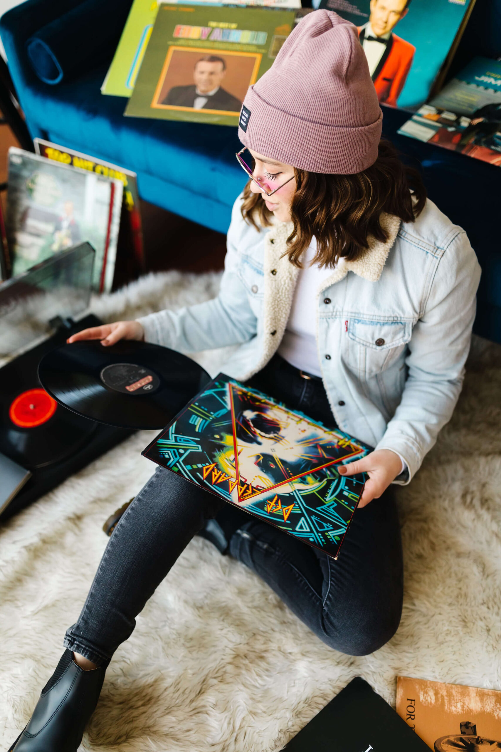 girl wearing pink beanie, blue jean jacket and neon pink sunglasses sitting in front of blue velvet couch putting a vinyl record album on a record player during senior studio photoshoot
