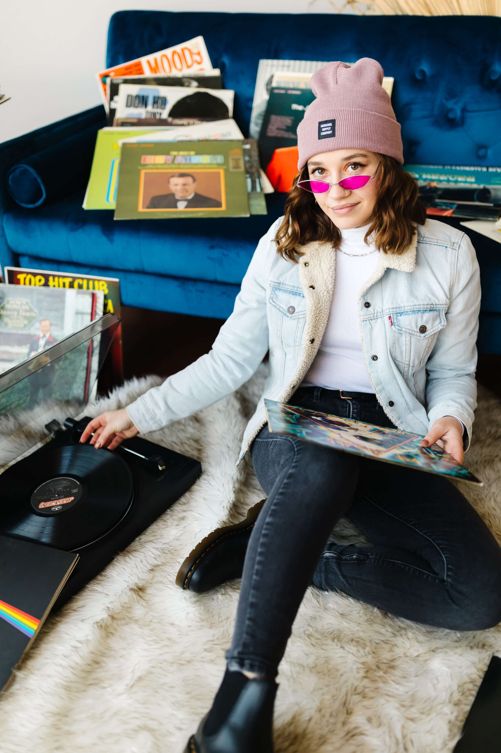 girl wearing pink beanie, blue jean jacket and neon pink sunglasses sitting in front of blue velvet couch putting a vinyl record album on a record player during senior studio photoshoot