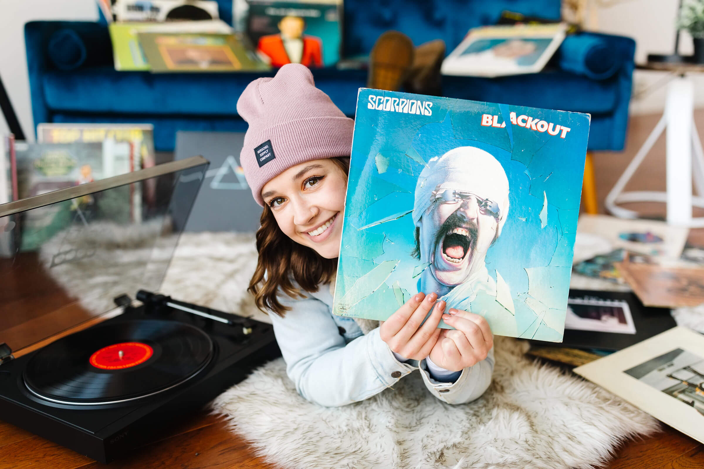 girl wearing pink beanie and blue jean jacket, laying on her stomach next to record player, holding vinyl record album in front of her face during senior studio photoshoot