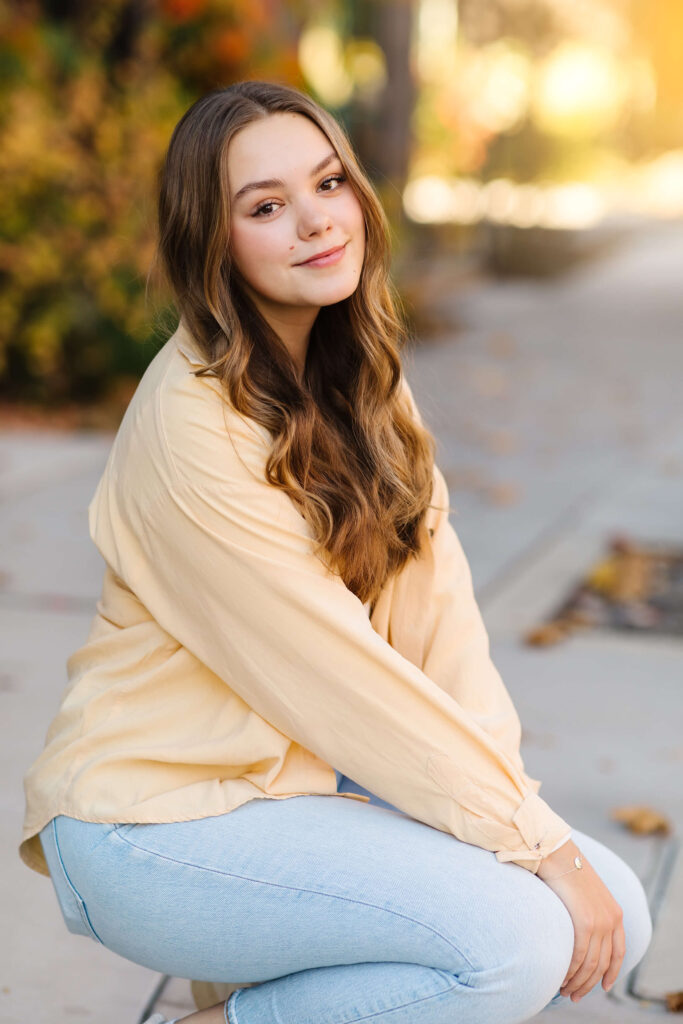 a girl in a yellow shacket kneeling down on sidewalk with colorful leaves during golden hour senior photos 