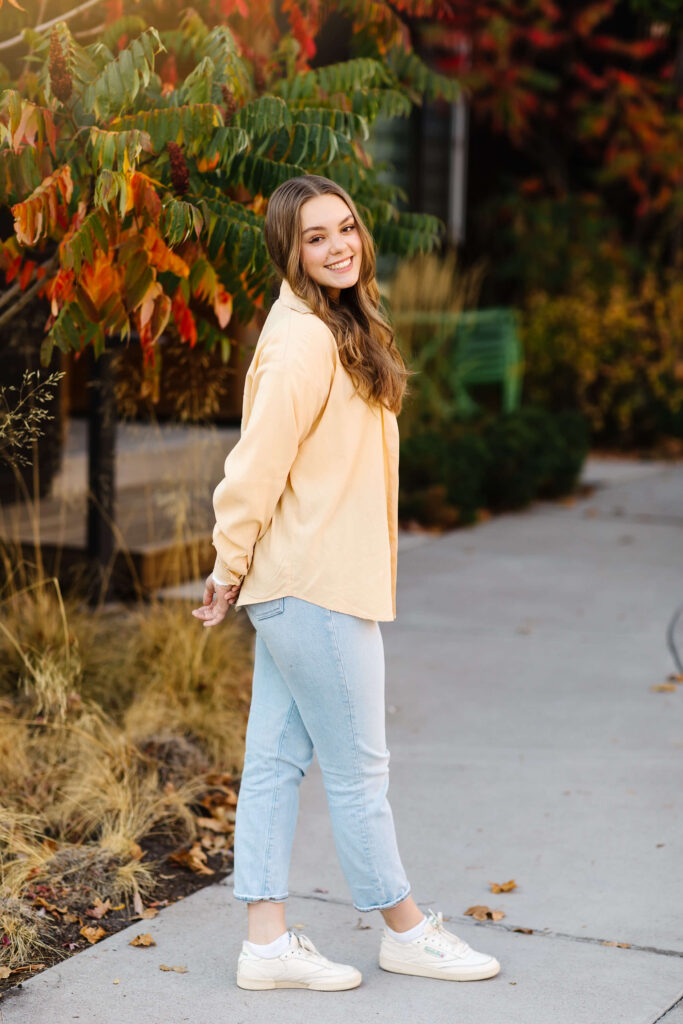 a girl in a yellow shacket standing on sidewalk with colorful leaves during golden hour senior photos 