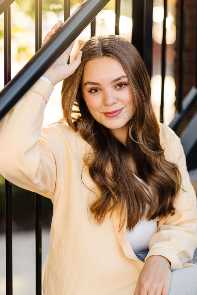 a girl in a yellow shacket smiling sitting on industrial black steel staircase during golden hour senior photos 