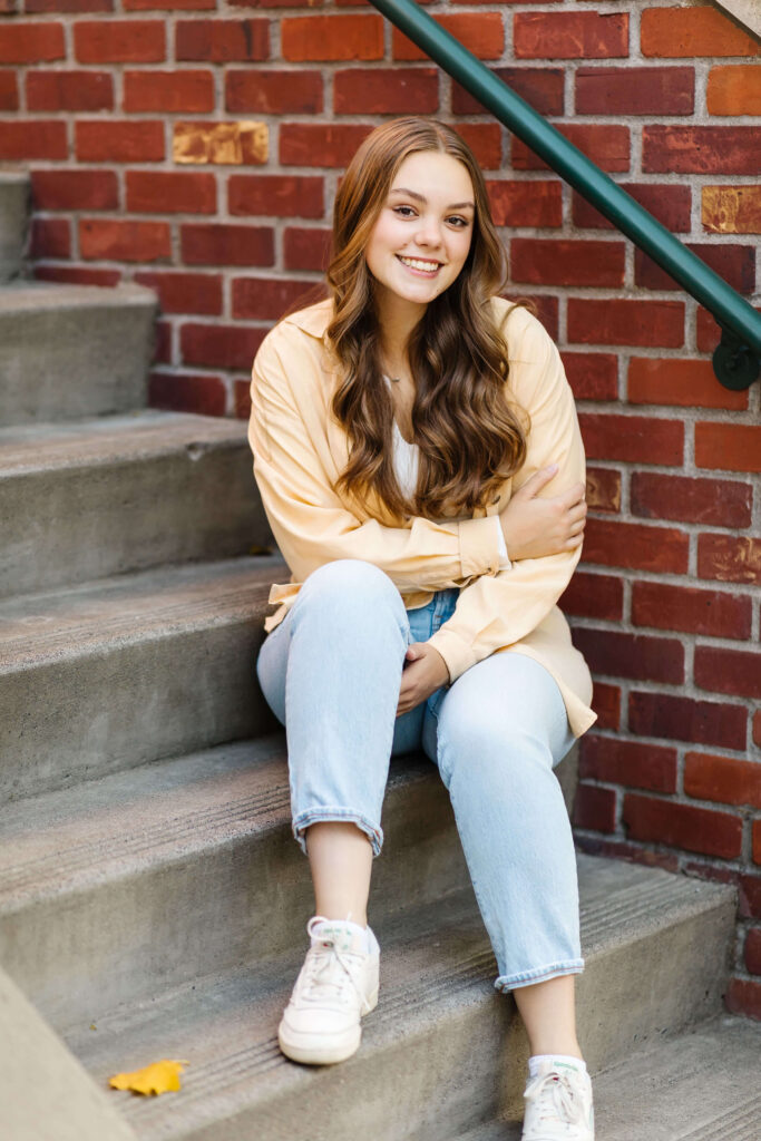 a girl in a yellow shacket sitting on cement staircase next to brick buildings smiling during golden hour senior photos 