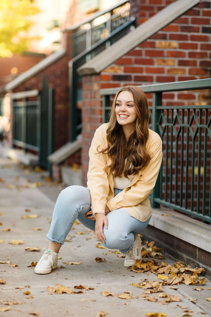 a girl in a yellow shacket sitting on cement staircase next to brick buildings during golden hour senior photos 