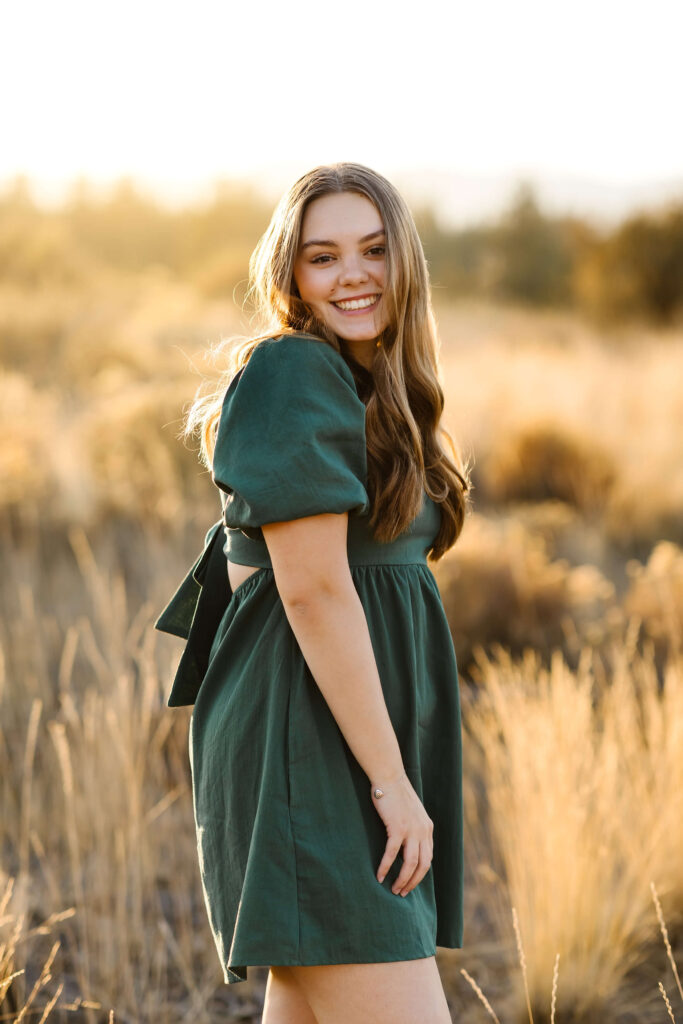 a girl with caramel color hair wearing green linen dress standing in tall grass field during golden hour senior photos session in bend oregon