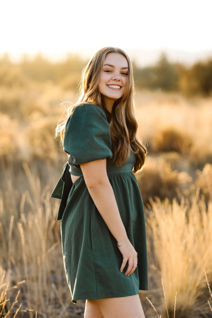 a girl with caramel color hair wearing green linen dress standing in tall grass field during golden hour senior photos session in bend oregon
