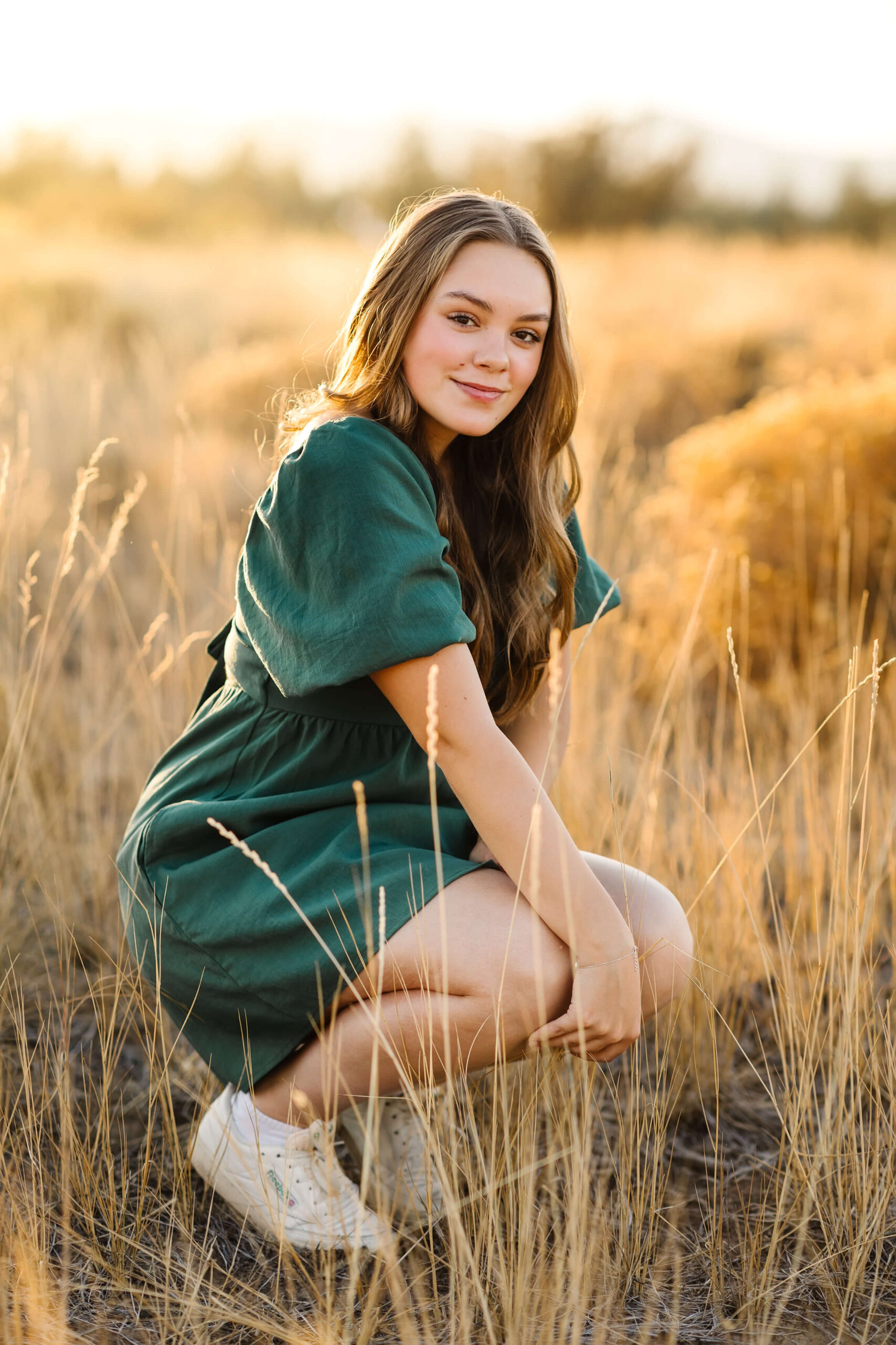 a girl with caramel color hair kneeling in tall grass field during golden hour senior photos session in bend oregon