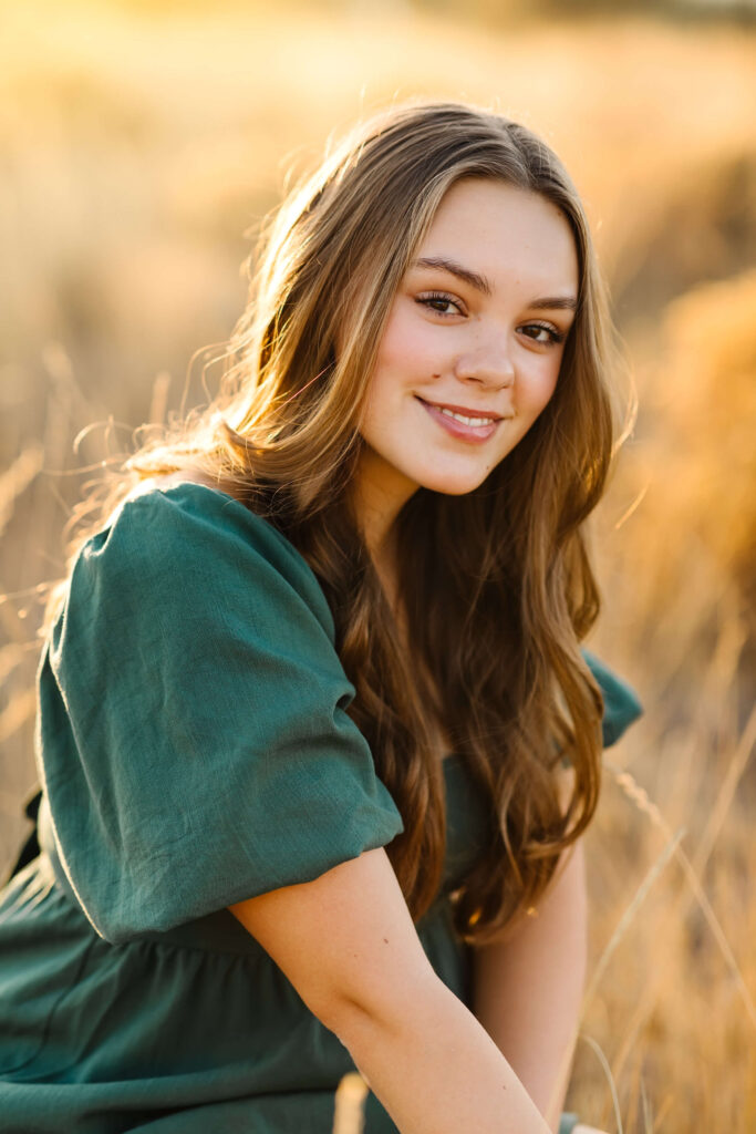 a girl with caramel color hair wearing green linen dress kneeling in tall grass field during golden hour senior photos session in bend oregon