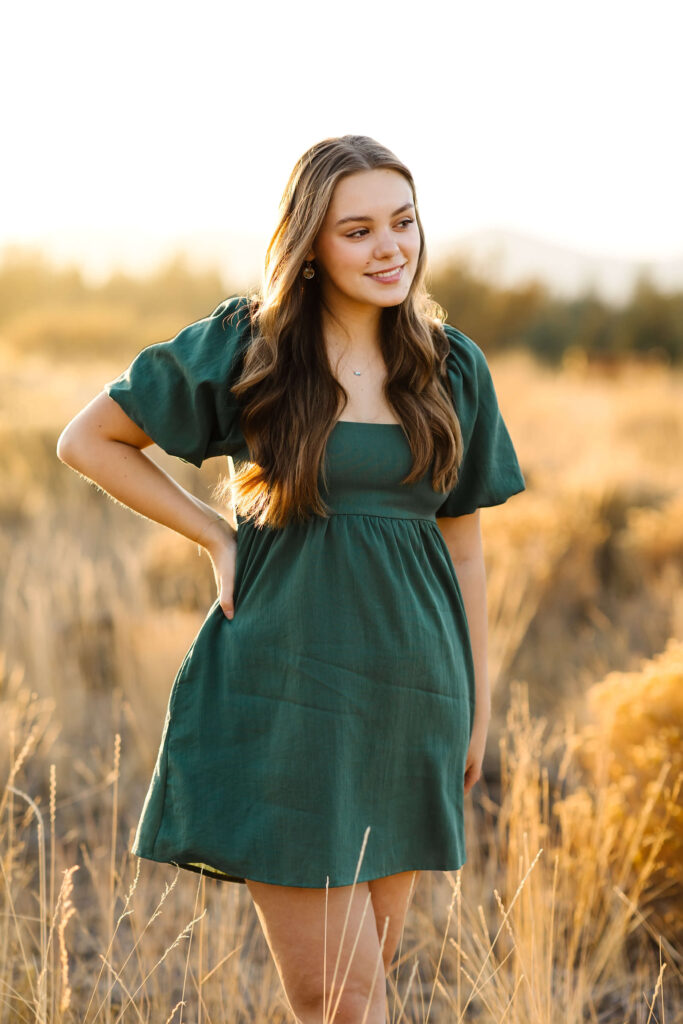 a girl with caramel color hair standing in a tall grass field in a green linen dress during golden hour senior photos session in bend oregon