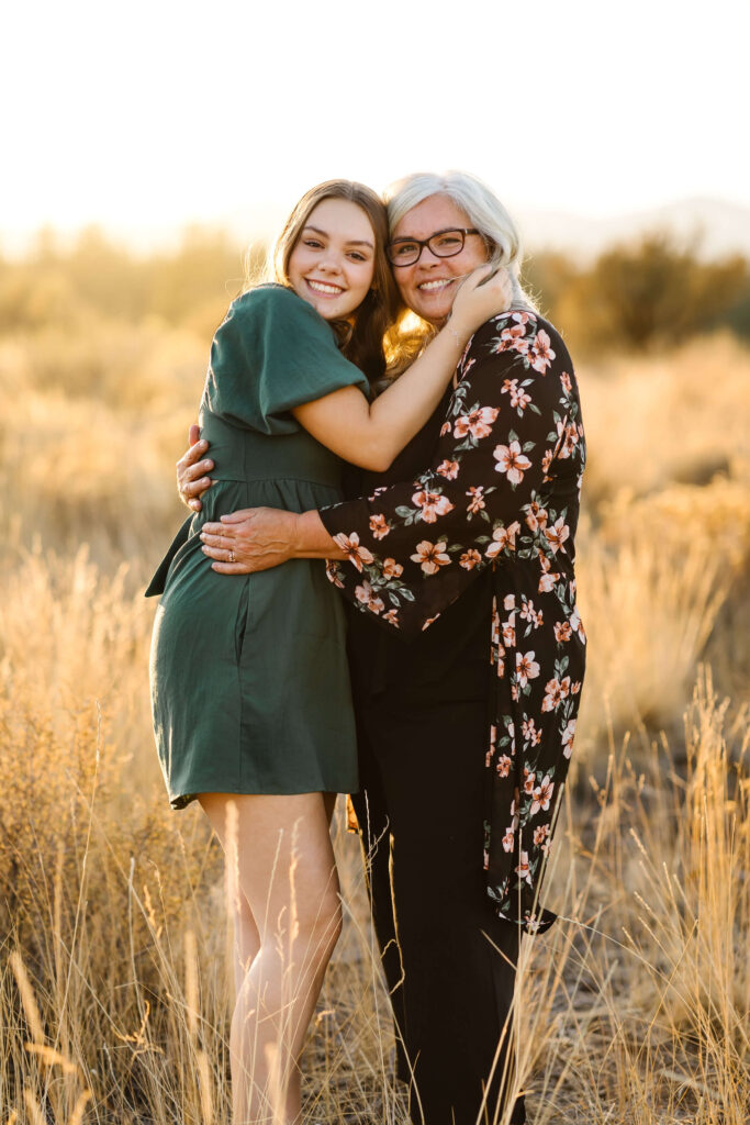 a girl wearing green linen dress hugging her mother in tall grass field during golden hour senior photos session in bend oregon