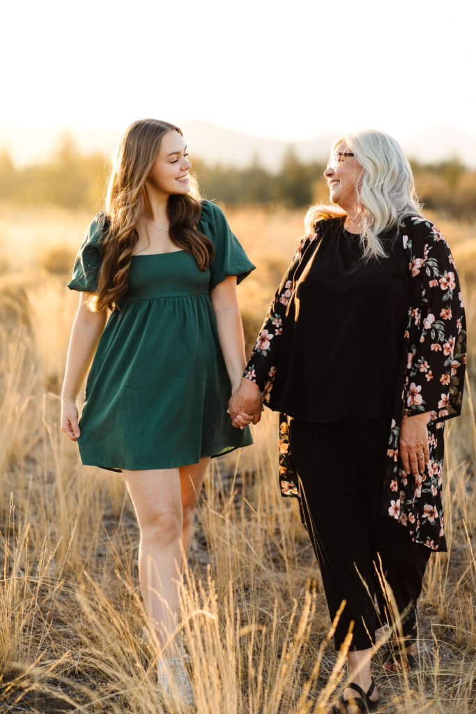 a girl wearing green linen dress holding hands and walking with her mother in tall grass field during golden hour senior photos session in bend oregon