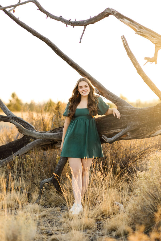 a girl with caramel color hair wearing green linen dress standing next to large log during golden hour senior photos session in bend oregon