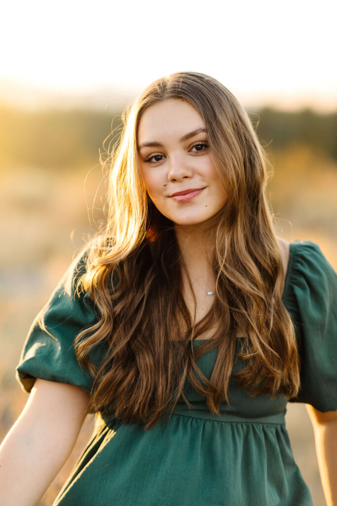 a girl with caramel color hair in a green linen dress standing in a tall grass field smiling during golden hour senior photos session in bend oregon